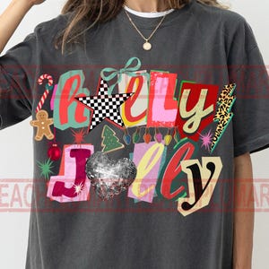 K&ouml;nnte beinhalten: Dunkelgraues T-Shirt mit einem farbenfrohen "Holly Jolly"-Grafik. Das Design umfasst einen Stern, ein Herz, einen Lebkuchenmann und einen Weihnachtsbaum. Der Text ist in verschiedenen Farben und Schriftarten gehalten, mit festlichem Thema.