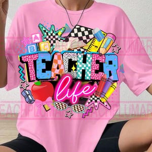 May include: Pink t-shirt with a colorful graphic design. The design includes the words "TEACHER life" in a mix of fonts and patterns, along with school-themed elements like an apple, crayons, and stars. The shirt is a casual, oversized fit.