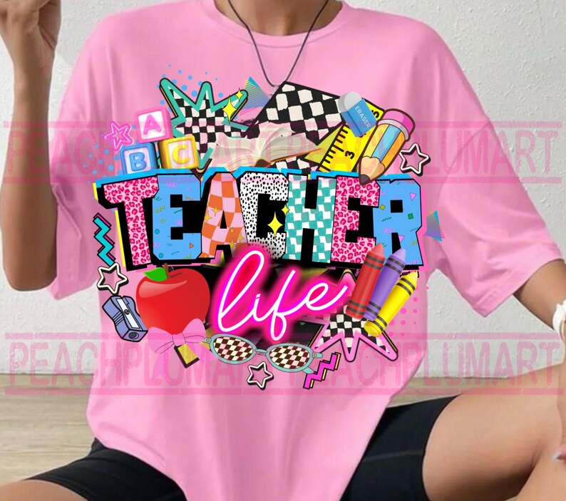 May include: Pink t-shirt with a colorful graphic design. The design includes the words "TEACHER life" in a mix of fonts and patterns, along with school-themed elements like an apple, crayons, and stars. The shirt is a casual, oversized fit.