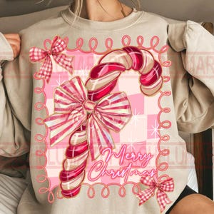 May include: A beige sweatshirt featuring a candy cane design with red and white stripes, adorned with pink bows and the text "Merry Christmas." The candy cane is set against a pink checkered background, with a decorative pink border.