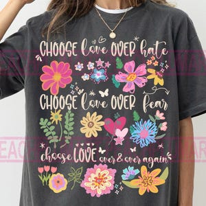 May include: Dark grey t-shirt with the words "CHOOSE love OVER hate", "CHOOSE love OVER fear", and "choose LOVE over & over again" in white script. The shirt features colourful floral and butterfly graphics.