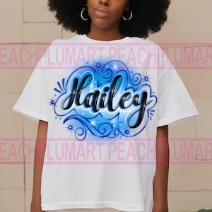 Peut inclure: T-shirt blanc avec un motif a&eacute;rographe bleu et noir repr&eacute;sentant le nom "Hailey" dans une police stylis&eacute;e avec des volutes et des &eacute;toiles.
