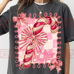 May include: Dark gray t-shirt featuring a candy cane design with red and white stripes, adorned with bows and the text "Merry Christmas." The design is set against a pink checkered background, creating a festive holiday theme.