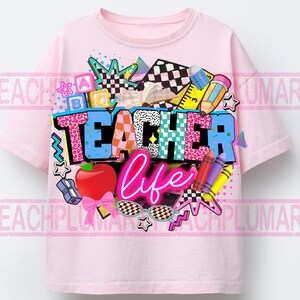 May include: A light pink t-shirt with a colorful graphic design. The design features the words "TEACHER life" in a mix of fonts and patterns, along with school-themed elements like an apple, pencils, and sunglasses. The shirt is a crew neck style.