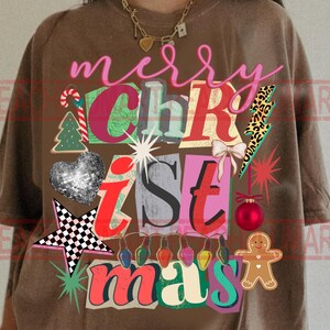 May include: Brown t-shirt with a colorful "Merry Christmas" graphic. The design includes a Christmas tree, heart, star, and gingerbread man. The text is in a mix of fonts and colors, with a festive holiday theme.