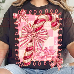 May include: Black t-shirt featuring a candy cane design with red and white stripes and a large pink bow. The design includes the text "Merry Christmas" in a cursive font, and a pink checkered background.