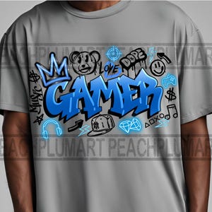 Op de afbeelding: Een grijs T-shirt met een blauw en zwart grafisch ontwerp met de tekst "Gamer". Het ontwerp bevat een kroon, een berenkop, een diamant, een noot, een bliksem, een koptelefoon, een controller en de woorden "Dope" en "Love".