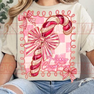 May include: A beige t-shirt featuring a candy cane design with a large bow and the words "Merry Christmas". The candy cane is red and white, set against a pink checkered background with a pink border.