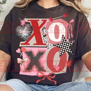 May include: A dark brown t-shirt featuring a Valentine's Day design with the letters "XO" in red. The design includes a disco ball heart, a checkered star, hearts, bows, and an arrow. The overall theme is love and romance.