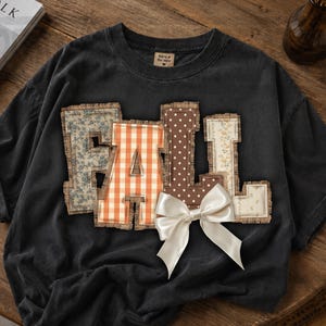 Könnte beinhalten: Ein anthrazitfarbenes T-Shirt mit dem Wort FALL in großen, aus Stoff applizierten Buchstaben mit ausgefransten Rändern. Jeder Buchstabe hat ein anderes Muster: blaues Blumenmuster, orangefarbenes Vichy-Karo, braune Punkte und cremefarbene Blumen.