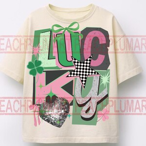 May include: Cream-colored t-shirt with the word "LUCKY" in a colorful, layered design. The design includes a green bow, a checkered star, a disco ball heart, and shamrocks. The letters are in pink, green, and gray.