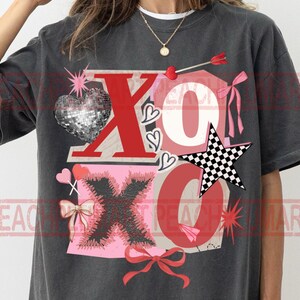 May include: Dark gray t-shirt with a graphic design featuring the letters "XO" in red and pink, a disco ball heart, a checkered star, and bow accents. The shirt has a vintage, distressed look.