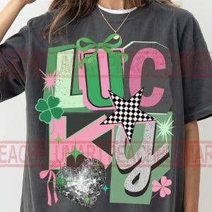 May include: Dark gray t-shirt with the word "LUCKY" in green, pink, and gray. The design includes a four-leaf clover, a disco ball heart, a checkered star, and a bow.