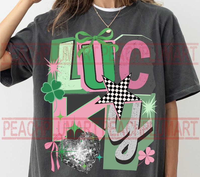 May include: Dark gray t-shirt with the word "LUCKY" in green, pink, and gray. The design includes a four-leaf clover, a disco ball heart, a checkered star, and a bow.