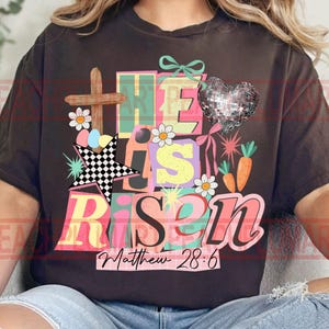 May include: Dark brown t-shirt with a colorful graphic that reads "He Is Risen" with a cross, star, and heart. The text "Matthew 28:6" is below the main text. The design includes daisies, carrots, and Easter eggs.