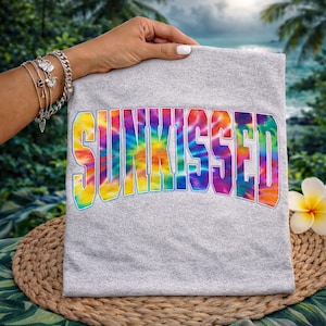 以下が含まれることがあります： タイダイデザインの「SUNKISSED」という文字がプリントされたグレーのTシャツ。 文字は太字でアーチ状になっており、青い縁取りがある。 熱帯の背景を背景に、手がTシャツを持っています。