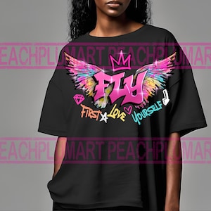 Może przedstawiać: Czarny T-shirt z grafiką w kolorze różowym, żółtym i niebieskim przedstawiającą skrzydła, koronę i napis "FLY". Grafika zawiera również tekst "First Love Yourself".