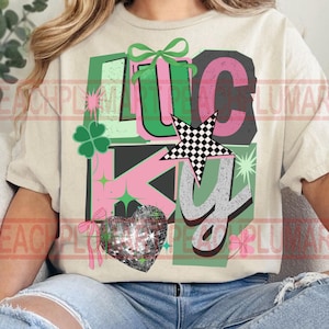 May include: Cream-colored t-shirt with the word "LUCKY" in a colorful design. The design includes a four-leaf clover, a checkered star, a disco ball heart, and bows. The letters are in green, pink, and gray.