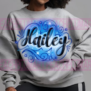 Peut inclure: Un sweat-shirt gris avec un motif a&eacute;rographe bleu et blanc repr&eacute;sentant le nom "Hailey" en lettres cursives noires.