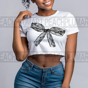 K&ouml;nnte beinhalten: Ein wei&szlig;es, bauchfreies T-Shirt mit einem schwarzen Spitzenbogen-Design. Das Shirt wird von einer Person mit dunkler Haut und kurzen, lockigen Haaren getragen. Die Person tr&auml;gt eine blaue Jeans.