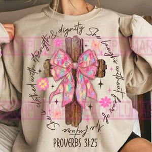 May include: A beige sweatshirt with a pink and white bow on a wooden cross. The text "She is clothed with strength & dignity She laughs without fear of the future Proverbs 31:25" is printed on the sweatshirt.