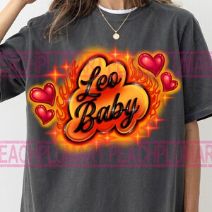 May include: Dark gray t-shirt with a vibrant graphic design. The design features the words "Leo Baby" in a stylized font, surrounded by flames, stars, and red heart shapes. The shirt is a casual wear item.