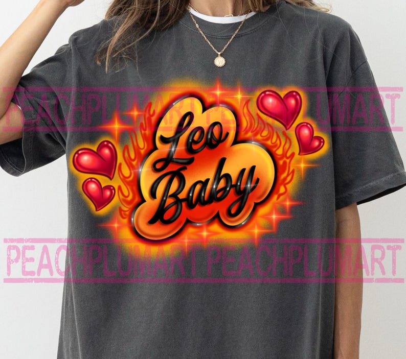 May include: Dark gray t-shirt with a vibrant graphic design. The design features the words "Leo Baby" in a stylized font, surrounded by flames, stars, and red heart shapes. The shirt is a casual wear item.