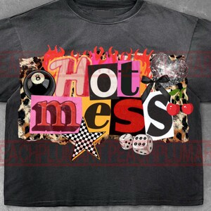 以下が含まれることがあります： 「Hot Mess」と書かれたカラフルなグラフィックが特徴の濃いグレーのTシャツ。グラフィックには、ヒョウ柄の背景、8ボール、ディスコボールのハート、さくらんぼ、サイコロ、市松模様の星が含まれています。