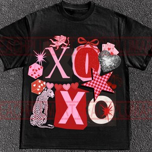 May include: Black t-shirt with a graphic design featuring the letters "XO" in pink and red, along with a leopard, dice, hearts, a star, and a cherub. The design has a vintage, distressed look.