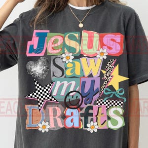 May include: Dark gray t-shirt with the text "Jesus Saw My Drafts" in colorful, block letter style. The design includes a disco ball heart, a checkered star, a smiley face, and other graphic elements.