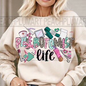 May include: A cream-colored sweatshirt with the text "Phlebotomist life" in pink, green, and black letters. The text is surrounded by various medical-themed illustrations, including a tourniquet, alcohol prep pad, bandages, and a syringe.