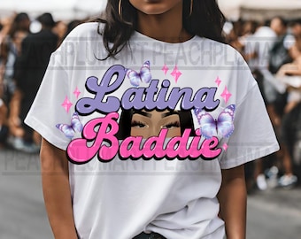 May include: White t-shirt with a graphic design featuring the text "Latina Baddie" in pink and purple lettering with a cartoon illustration of a woman's eyes and eyelashes. The design also includes butterflies and stars.