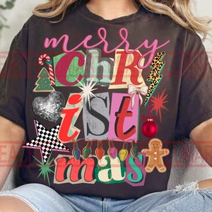 Può includere: T-shirt marrone scuro con una grafica natalizia colorata. Il design include le parole "merry Christmas" in rosa e vari elementi festivi come un bastoncino di zucchero, un albero di Natale e un omino di pan di zenzero.