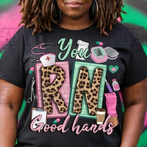 Może przedstawiać: Czarny T-shirt z nadrukiem w kolorze r&oacute;żowym, zielonym i w panterkę z napisem "You RN Good Hands". Nadruk zawiera stetoskop, czapkę pielęgniarki, pudełko z tabletkami, mankiet do pomiaru ciśnienia krwi, strzykawkę i kubek do kawy.