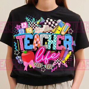 May include: Black t-shirt with a colorful "Teacher Life" graphic. The design includes an apple, pencils, crayons, and the words "Teacher Life" in a neon pink font. The word "Teacher" is in a mix of patterns and colors.