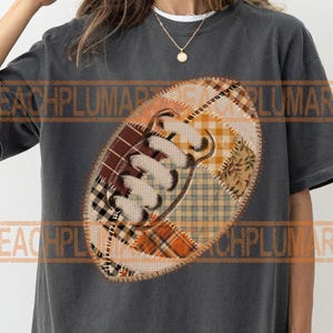 May include: Dark gray t-shirt featuring a football design. The football is made of patchwork fabric in shades of brown, orange, and cream. The design has a stitched outline and a white lace detail.