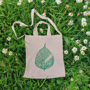 May include: A white tote bag featuring a green leaf design and the text "سَلامَتِكَ". The bag is resting on a bed of green grass and white daisies.