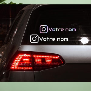 Peut inclure: Une voiture noire avec un autocollant sur la lunette arrière qui dit "Votre nom" avec une icône Instagram. L'autocollant est en blanc et rose.