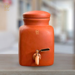May include: A terracotta water dispenser with a lid and a brass tap. The dispenser has a rounded top and a square base. The Mittify logo is visible on the front. It is designed to hold and dispense water.