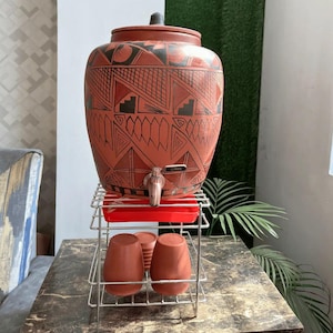 May include: A terracotta water dispenser with a lid and tap, decorated with black geometric patterns. It stands on a metal stand with a red tray, alongside two matching terracotta cups. The dispenser is designed for serving drinks.
