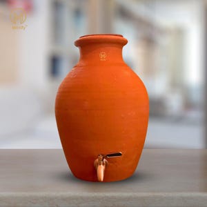 Könnte beinhalten: Ein Terrakotta-Wasserdispenser mit einem Kupferhahn. Der Dispenser ist im traditionellen Stil mit einem abgerundeten Körper und einer breiten Öffnung. Der Dispenser steht auf einer hellbraunen Oberfläche.