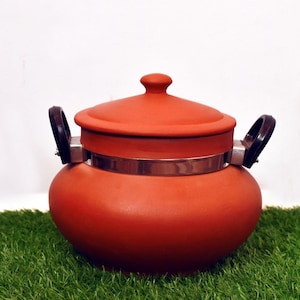 Puede incluir: Una olla de cocina de arcilla de terracota con tapa y dos asas negras. La olla está asegurada con una banda de metal plateado. La olla está sobre un lecho de césped artificial verde.