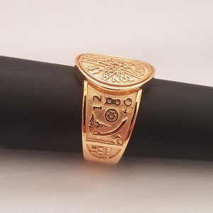 Può includere: Un anello d'oro con incisioni e simboli intricati, tra cui numeri e una stella. L'anello è su una superficie nera.