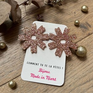 May include: A pair of glittery rose-gold flower-shaped earrings. The earrings are displayed on a white card with French text, "COMMENT TU TE LA PÊTES !" and "Bijoux Made in Niort". The background includes wooden planks, dried leaves, and gold decorative spheres.