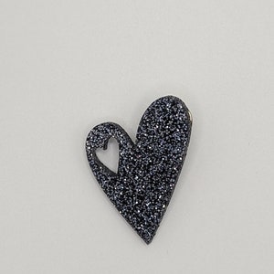 May include: A glittery, dark gray heart-shaped brooch with a smaller heart cut out. The brooch has a metallic sheen and is set against a plain white background. The heart is a decorative accessory.