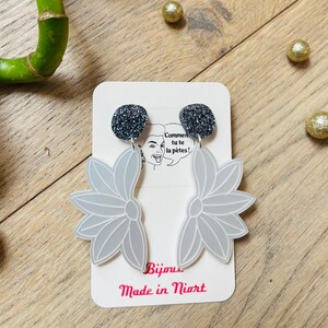 Peut inclure: Boucles d'oreilles grises en forme de fleur avec des clous noirs scintillants. Les boucles d'oreilles sont présentées sur une carte blanche avec du texte français et les mots "Bijoux Made in Niort". Le fond est une surface en bois brun clair.