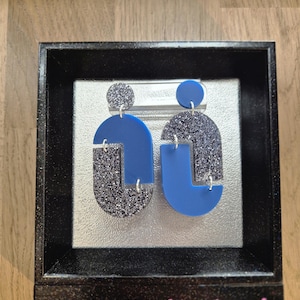 Könnte beinhalten: Ein Paar geometrischer Ohrringe in Blau und silbernem Glitzer, präsentiert in einem schwarzen Rahmen. Die Ohrringe haben ein modernes Design mit einfarbigen blauen und glitzernden silbernen Abschnitten. Der Text "Boucles d'oreilles du mois" ist sichtbar.