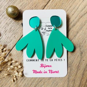 May include: Turquoise and teal statement earrings with a leaf-like design. The earrings are attached to a white card with French text and the words "Bijoux Made in Niort". The earrings are displayed on a wooden surface.