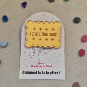 Peut inclure: Épinglette dorée Petite Nantaise sur une carte blanche. L'épinglette a la forme d'un biscuit avec le texte "PETITE NANTAISE". La carte porte également le texte "Bijoux Handmade in Niort" et "Comment tu te la pètes !"