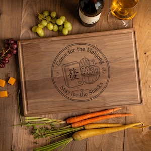 May include: A rectangular wooden cutting board featuring a laser-engraved circular design with a mahjong tile, a bowl of snacks, and the text "Come for the Mahjong Stay for the Snacks" surrounded by decorative lines and small heart icons.